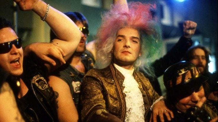 Still from the Rock Me Amadeus music video with Falco dressed as Mozart with a multi-color wig sitting with a group of bikers.