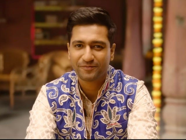 A still from Hindi film, ‘The Great Indian Family’. Bhajan Kumar aka Ved Vyas played by Vicky Kaushal is smiling. It is a bust shot with him wearing what looks like a kurta. He has a blue jacket with ornate golden embroidery.