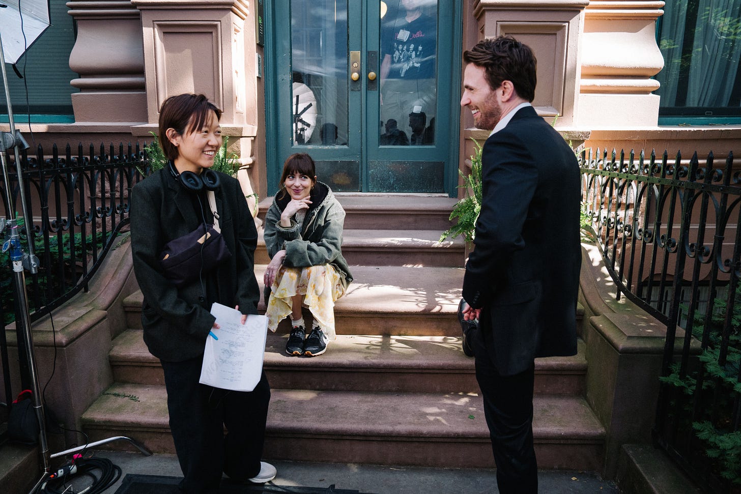Director Celine Song with actors Dakota Johnson and Chris Evans during the filming of Materialists. Song wears a set of headphones and is holding a script. She is chatting with Evans, while Johnson sits on a stoop, smiling at Evans. (A24) Director Celine Song with actors Dakota Johnson and Chris Evans during the filming of Materialists. Song wears a set of headphones and is holding a script. She is chatting with Evans, while Johnson sits on a stoop, smiling at Evans. (A24)