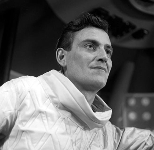 A young white man with dark hair. He wears a spacesuit without a helmet or breathing apparatus, and is within a space rocket.