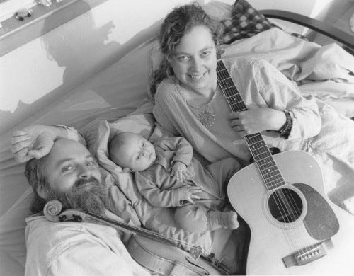 Fiddler and guitarist lying on bed with their new baby and instruments.