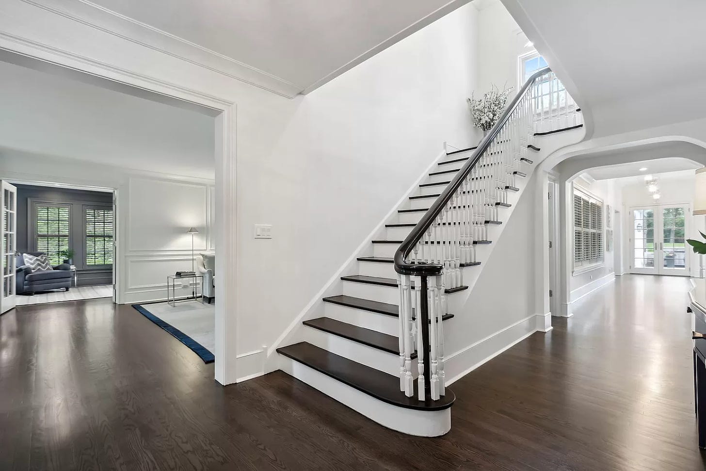 The inside of the house. Dark wood floors, the main staircase, and the walls are all white. This view allows you to see through to the living room and office, and everything is white. One far room has dark gray walls but wow, there is no character left here.