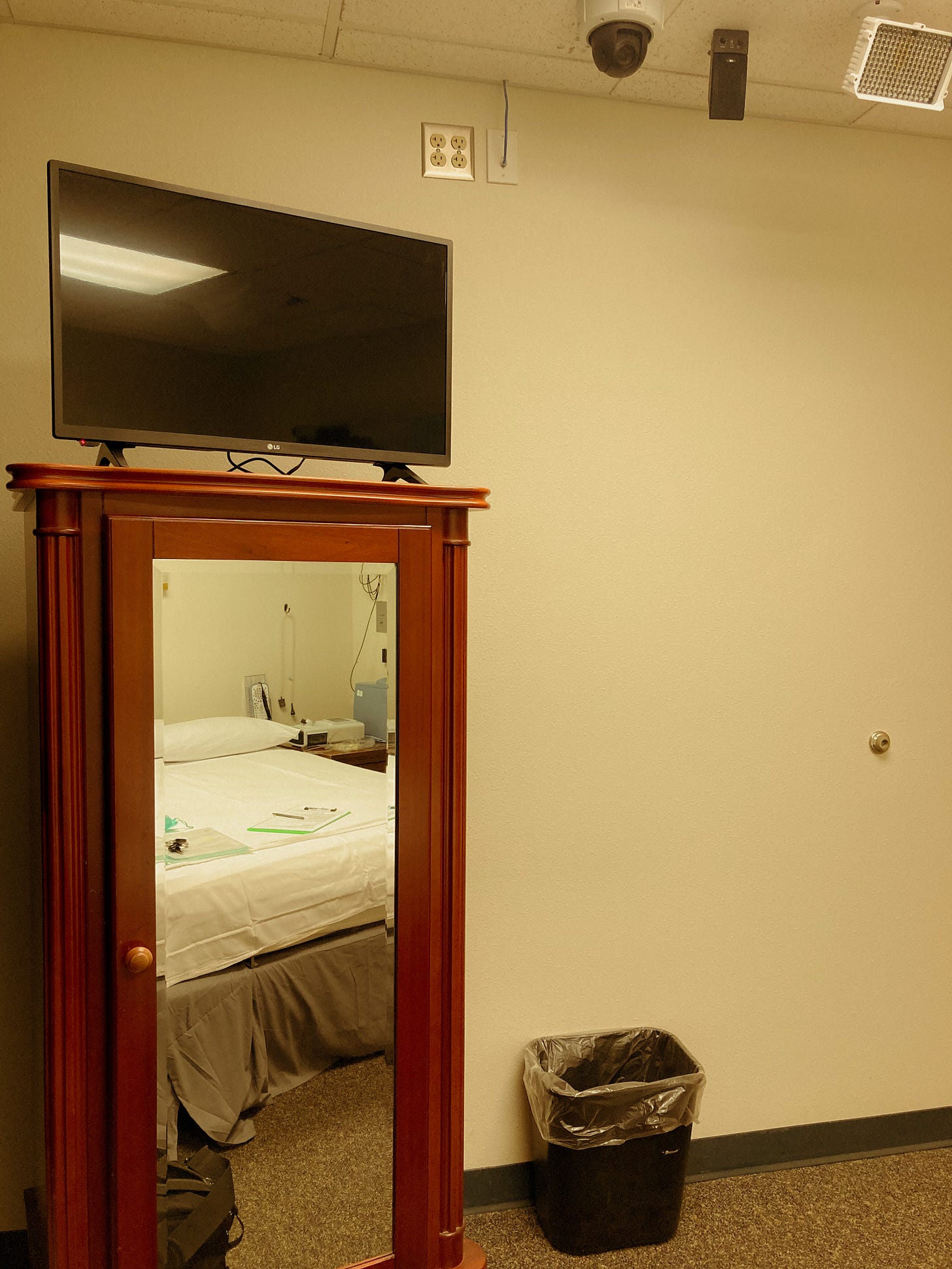 A white room with institutional ceiling tiles, with a mirror on an armoire reflecting a bed with white sheets and machines next to it.