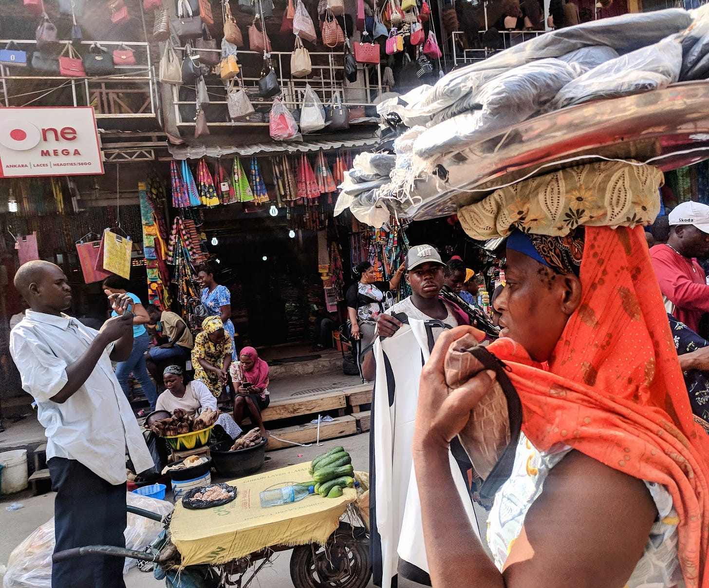 A man shields his phone to read it in the sun, and a woman with a bright scarf walks by with packages on her head. They are surrounded by a busy, colorful market, with people sitting on a stoop and hanging purses and cloths for sale.