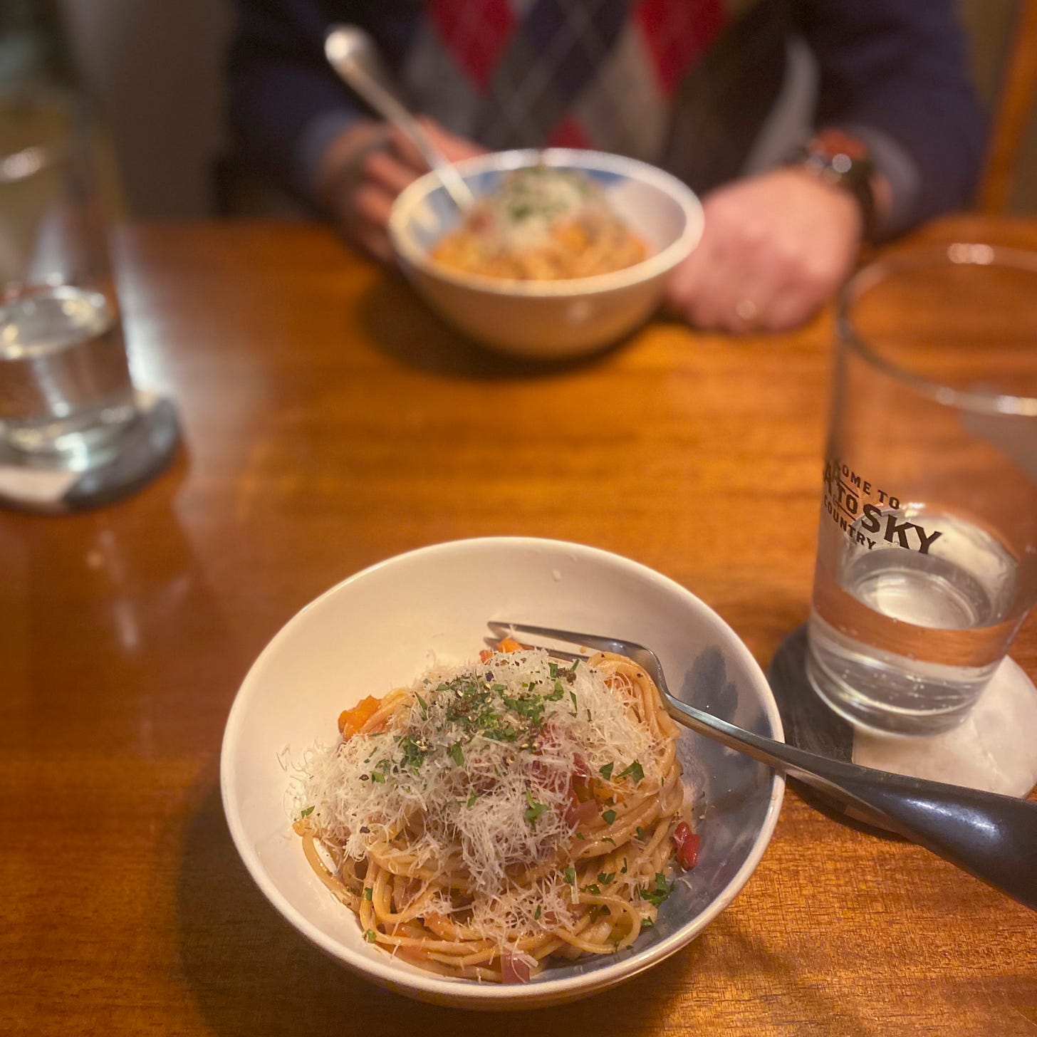 A white bowl of linguine in the sauce described above, with a hefty pile of shredded pecorino on top, as well as ground pepper and chopped parsley. Jeff is in the background with a bowl of the same, and two glasses of water sit on coasters next to the bowls.
