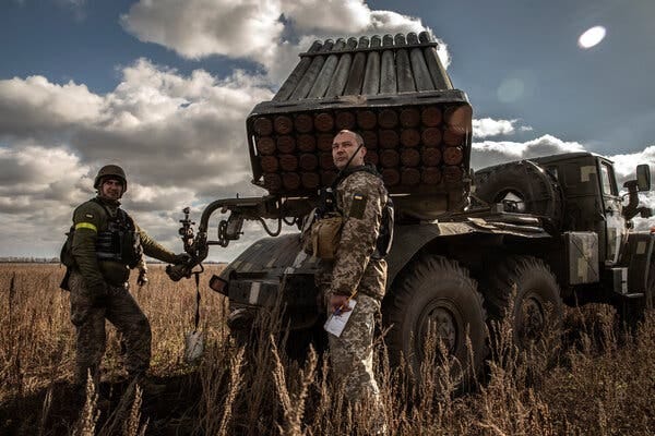 Two men in military gear standing next to rocket launcher.