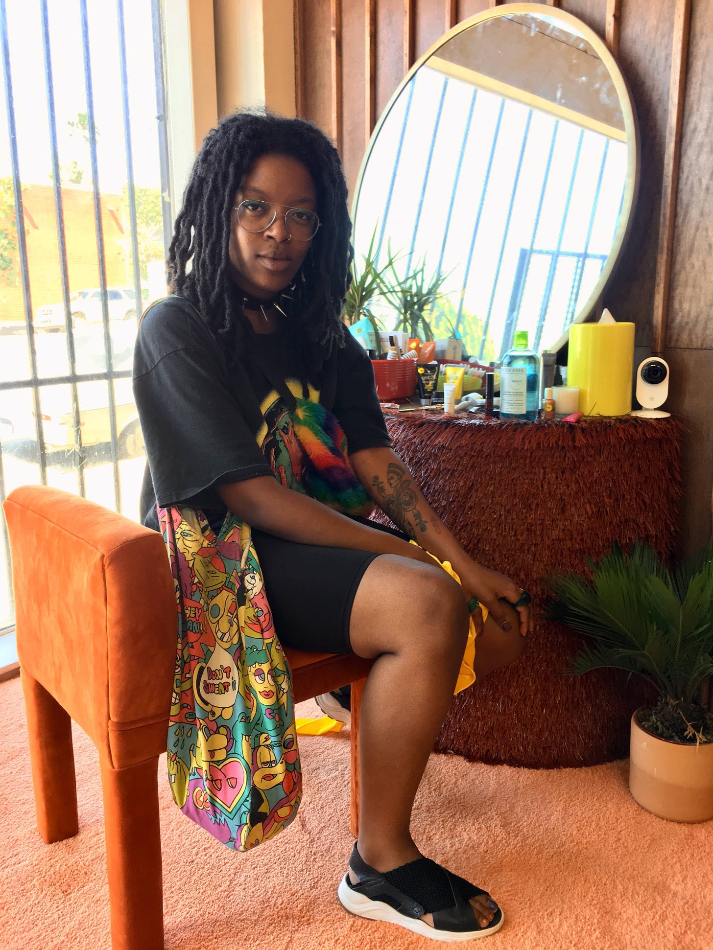 A young Black woman sits at a vanity inside a shop. she poses while wearing black clothing, aviator glasses, a spiked necklace, and two colorful bags.