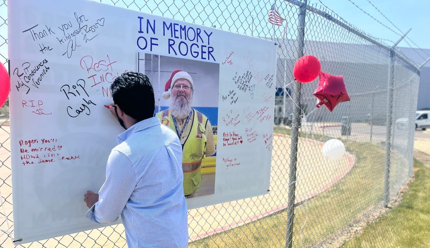 A man stands in front of a white poster with signatures reading "rest in peace" and "in memory of roger" that is hanging on a tall wire fence