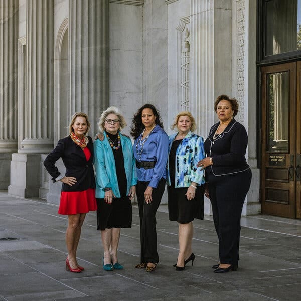 Five women stand in a row outside an ornate, white building.