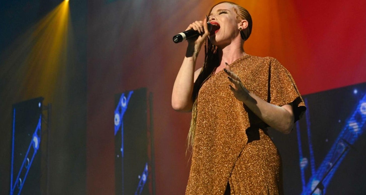 A femme singer with albinism is singing into a mic on a large stage. She wears a regal gold dress, red lipstick, and braids.