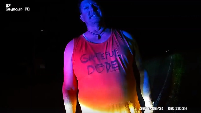 Police body cam stillshot of state Rep. Jim Lucas (R-Indiana) wearing a sleeveless shirt with the slogan 'Grateful Dude.'  The 'u' is a big smiley face with the eyes forming an umlaut. the image is lit only by police cruiser lights so Lucas's shirt appears red while his face is shaded blue.