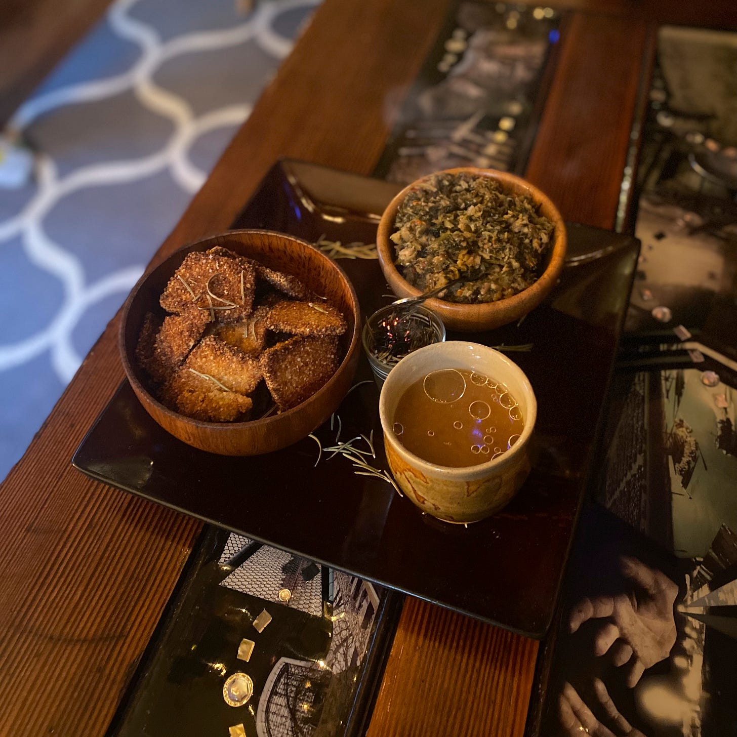 A square black plate with two small wooden bowls, a cup of broth with oil floated on top, and a small glass cup of incense. The bowl in the foreground holds square quinoa crackers, and the other bowl holds chutney with a spoon in it. The plate is dusted with rosemary.