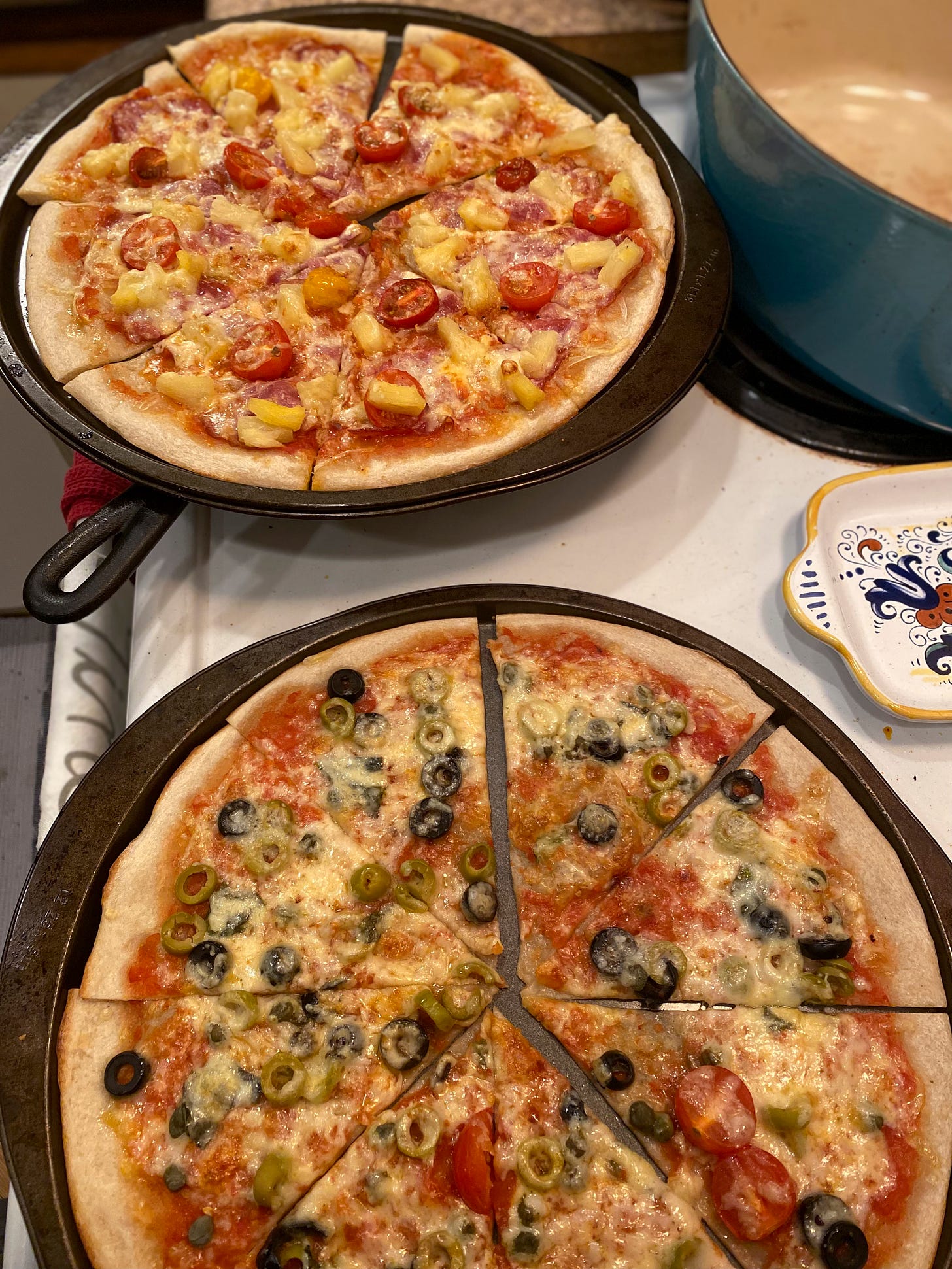 Two pizzas in their pans on the stove, sliced into eight pieces each. The one in the foreground has two kinds of olives, capers, and grape tomatoes, and in the background is one with capocollo, pineapple, and the same kind of tomatoes. Both are lightly covered in melted cheese that has browned in places.