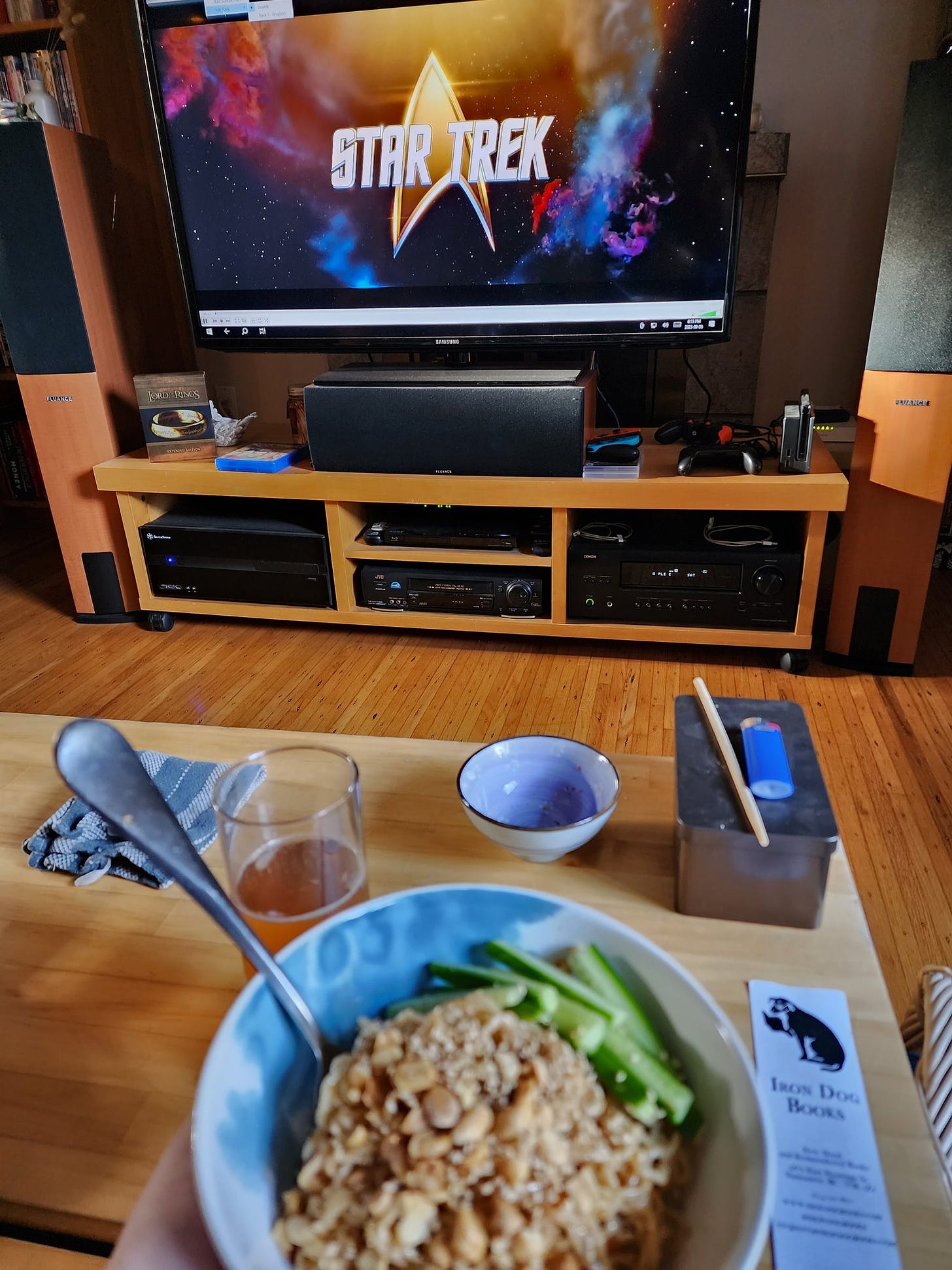 In the foreground, blurred, is a bowl of noodles in the sauce described above, topped with peanuts and with cucumber matchsticks on one side. The coffee table is visible behind it, with a few random things on it, and in focus in the background is the tv with the Star Trek title card.