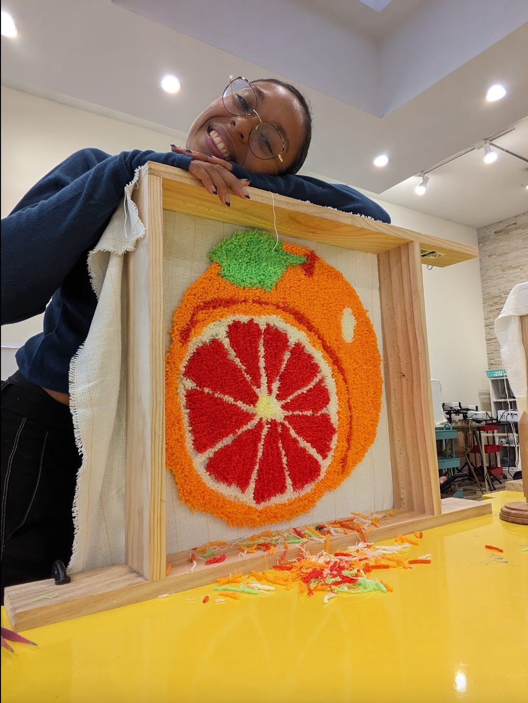 Nathalie is resting her arms and head atop her tufted rug panel. She has a big smile on her face, proud of the work she created. Her tufted rug is of a bright orange and red grapefruit.