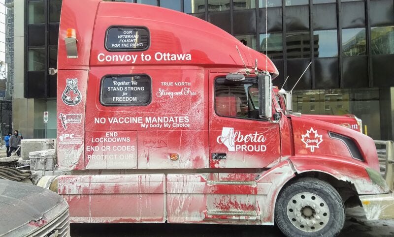 A dusty red truck cab, photographed from the side, covered in slogans for the Freedom Convoy. Slogans include "End lockdowns, end QR codes, protect our children," "No vaccine mandates; my body my choice," "True North, strong and free," and "Our veterans fought for the right!"