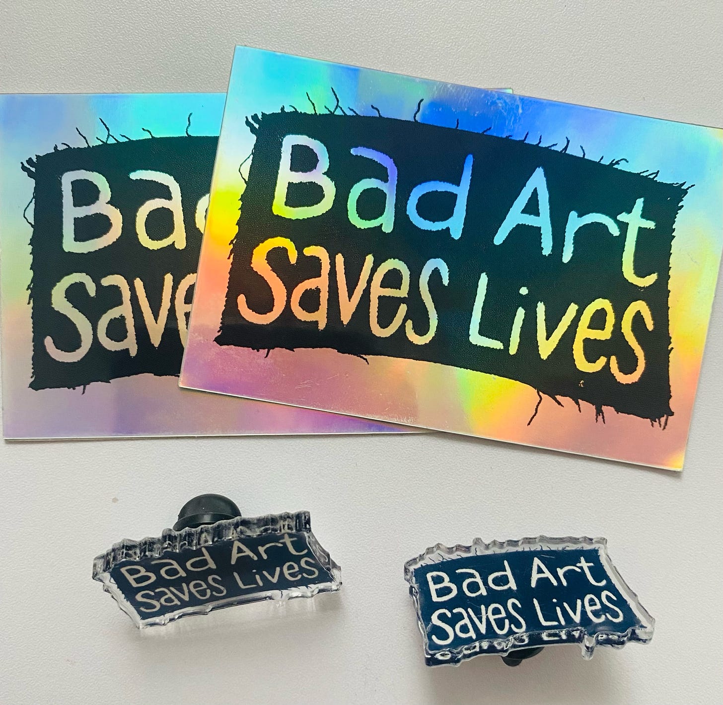 An image of two holographic "bad art saves lives" stickers and two black "bad art saves lives" pins.
