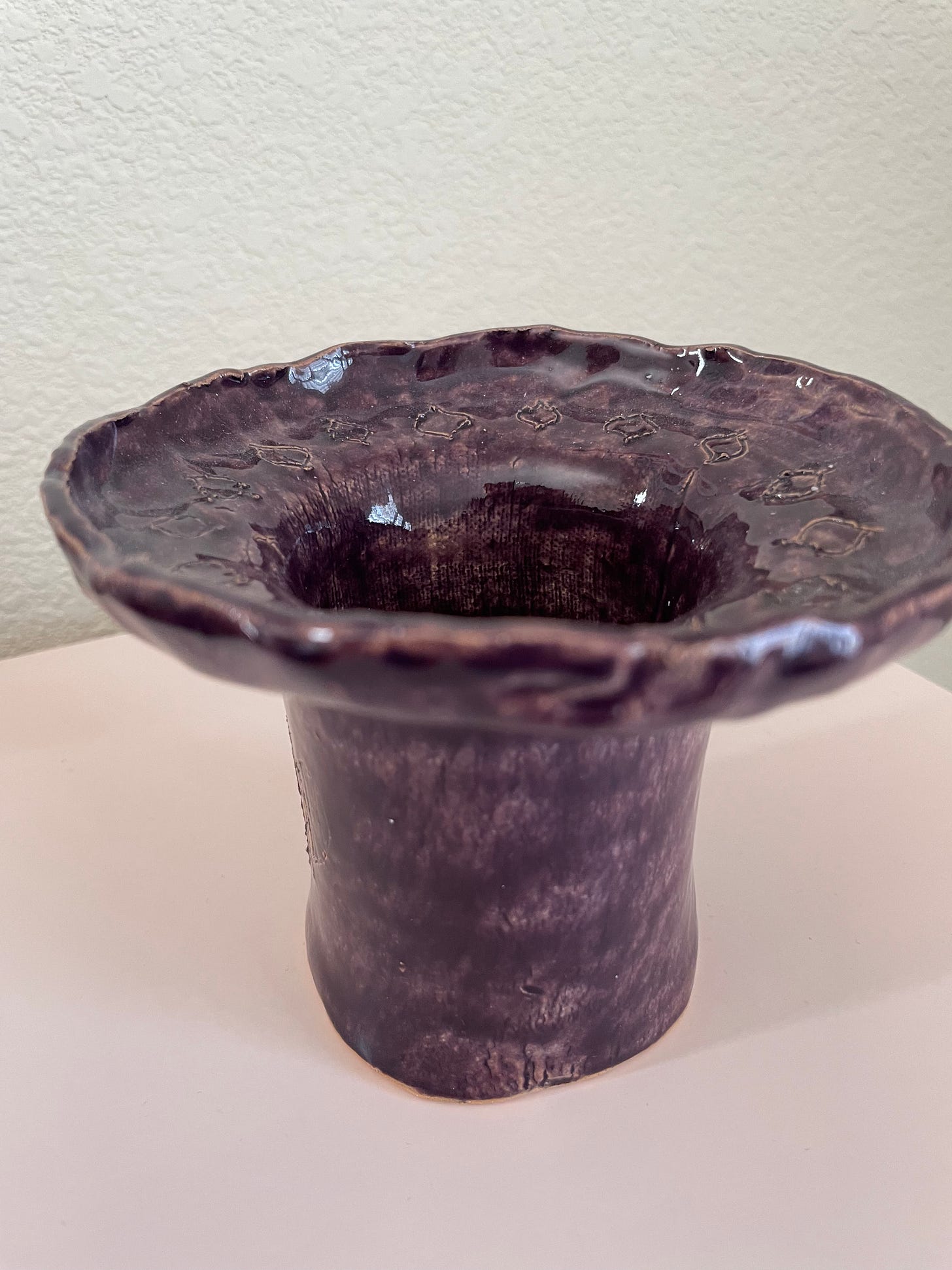 A dark purple vase-like glazed clay piece. It has little designs etched in and is atop a light pink box with a white wall in the background.