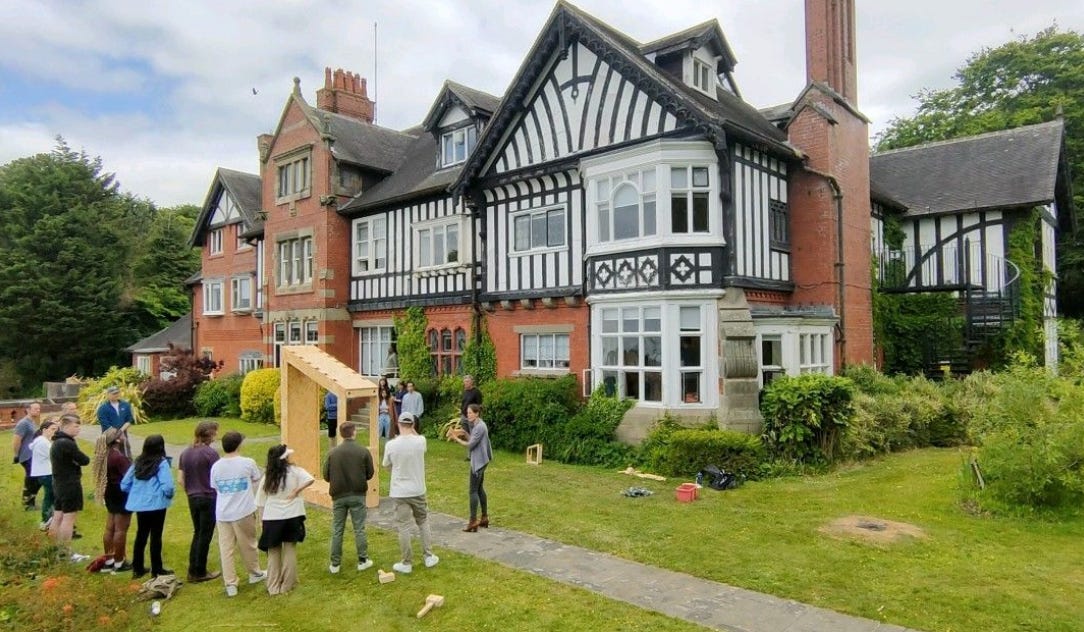 A group of students look at a model WikiHouse structure built in front of a larger Tudor style building