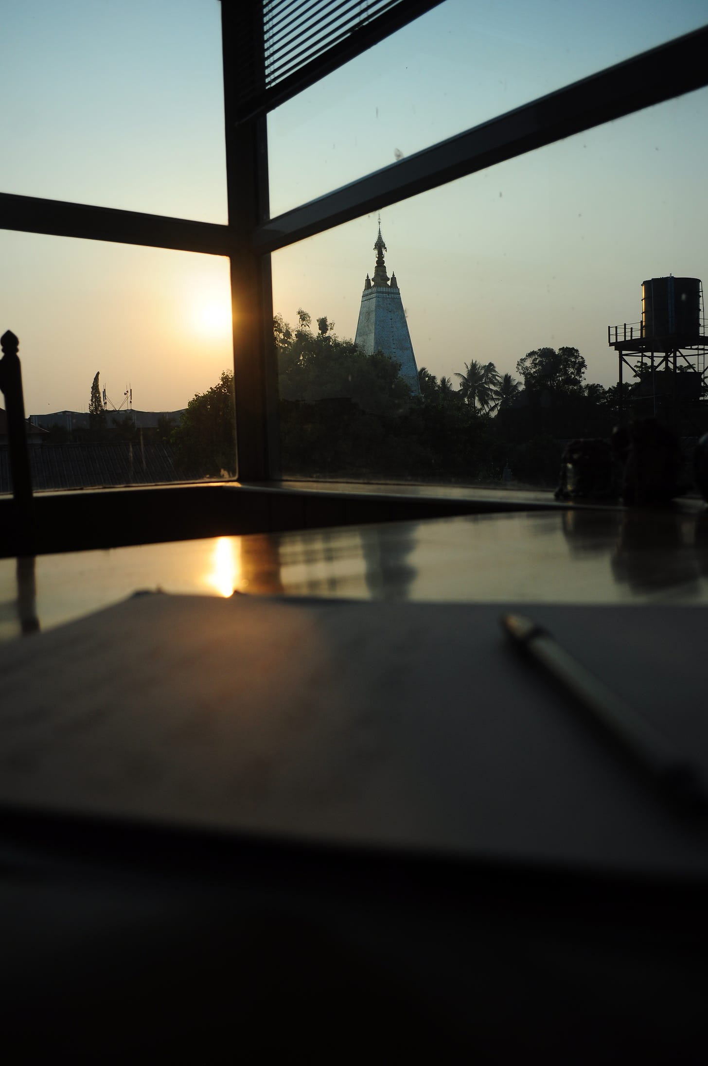 Out the window you can see the sunset, a yellow colour and the pyramid shape of a pagoda which is covered is mosaic mirror pieces. You can also see some trees and a water tank.