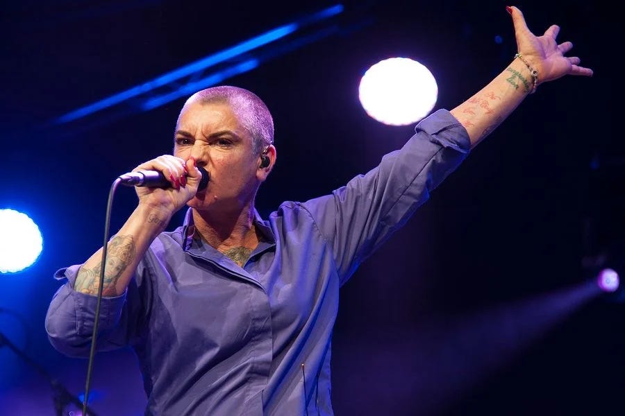 A white woman with a shaved head, greying at the temples, is singing into a microphone with passion, her left arm lifted up and hand extended in gesture.