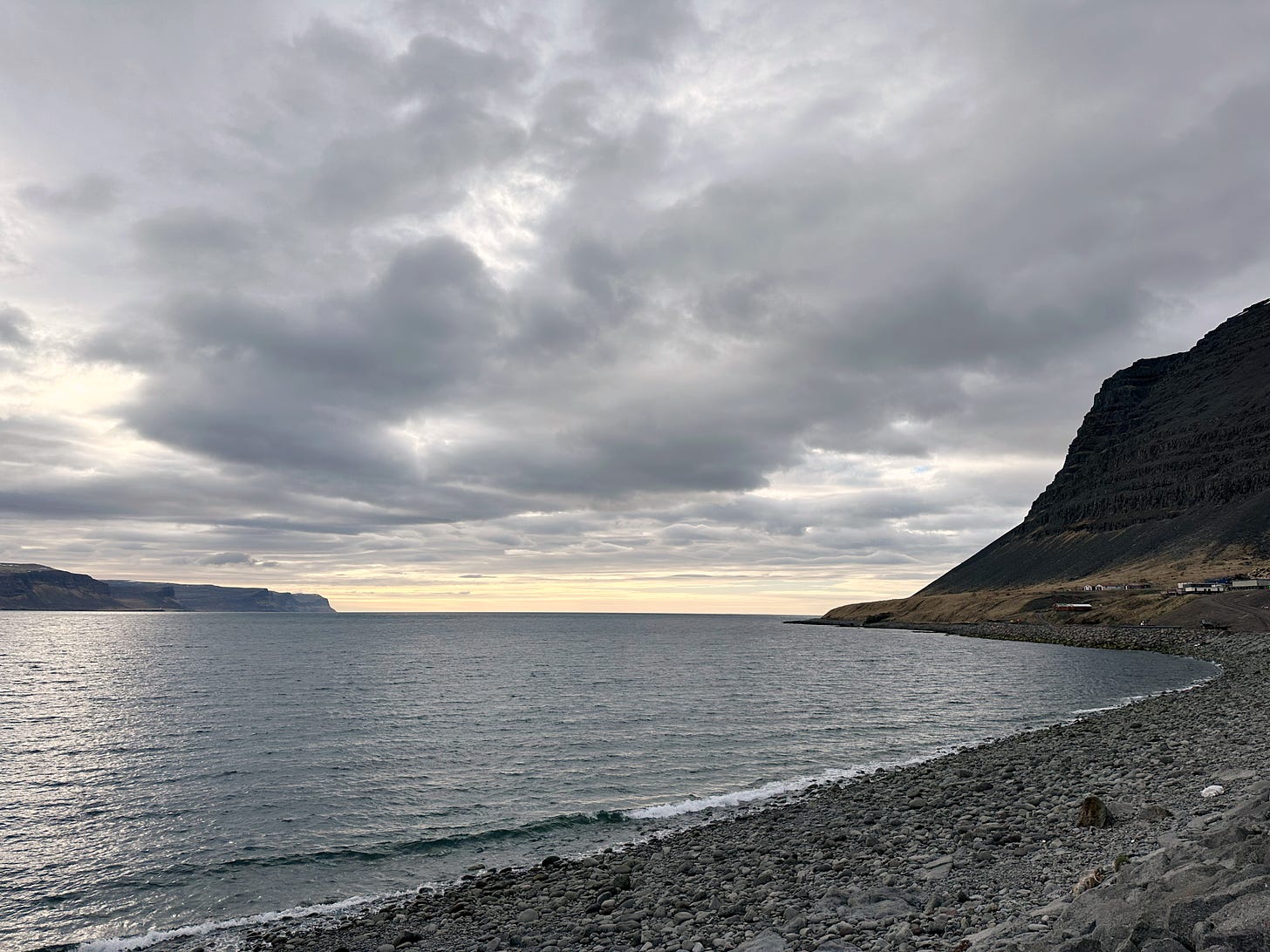 The view from the beach looking out towards the open sea.  High hillsides on the right with lower land on the left.  The sky is cloudy with the beginnings of sunset colors and the rocky beach curves to the right.