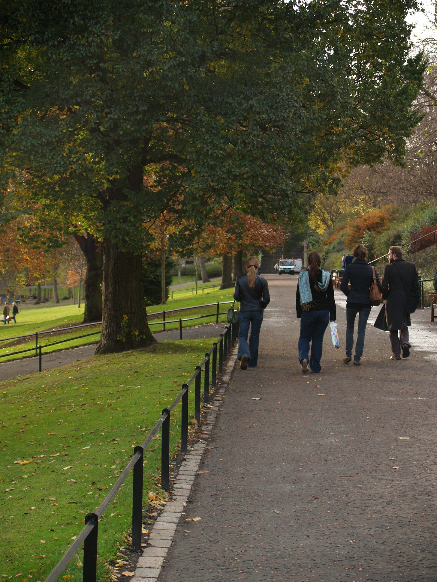 A line of people walking through a lush green park filled with grass and trees. The woman in the centre has a long golden-brown ponytail and is wearing a black leather jacket and jeans, and the woman to her right has dark hair and wears a blue scarf over a black pea coat. 