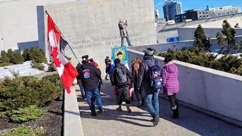 Several people, one carrying a flag, walk along a path to a large concrete monument.