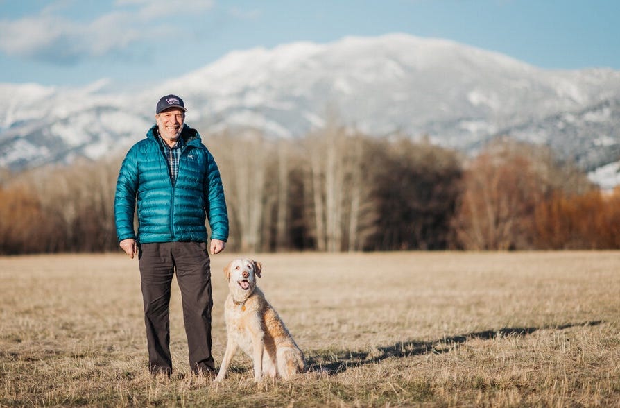 man in puffy with dog in field and mtns in background