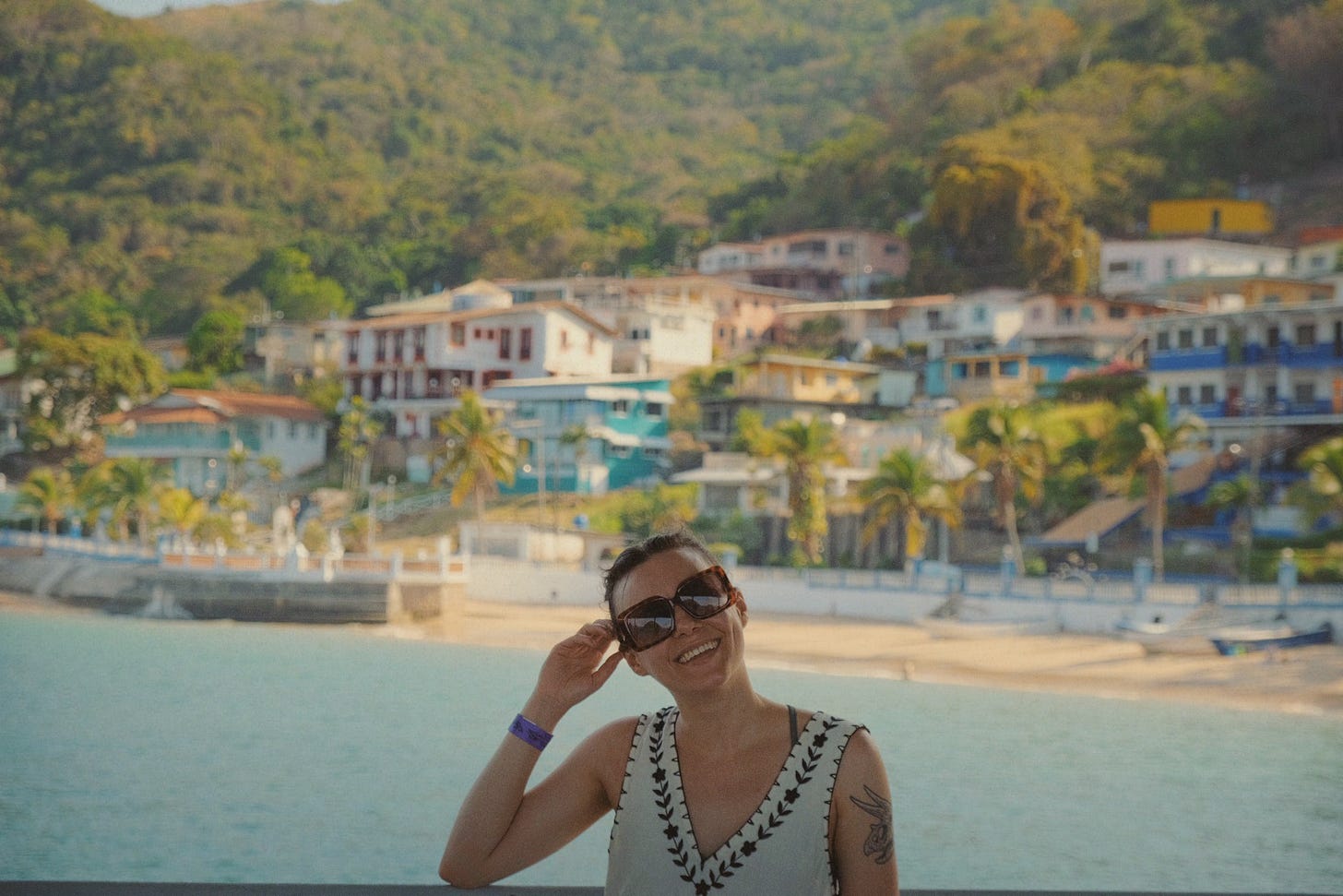 Leah in sunglasses and a sundress smiling at the camera, with the sun shining on the beach, boats, and hotels in the background.