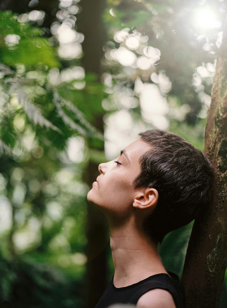 Woman with eyes closed meditates in forest near a tree.