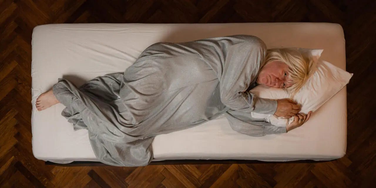 A man in a kaftan and blonde wig lays on a bed.
