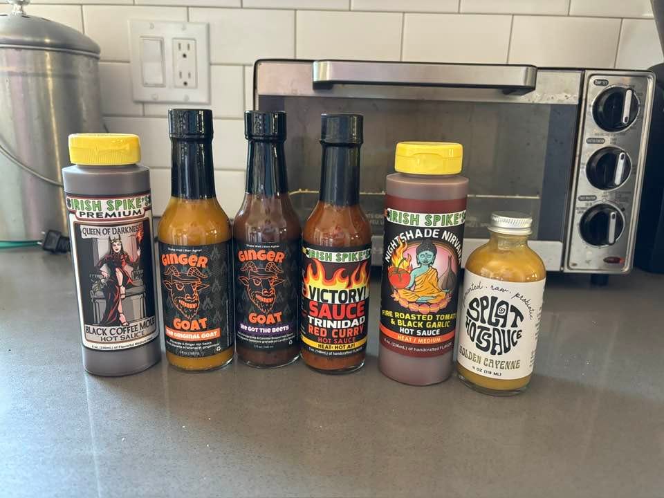 Six bottles of hot sauces lined up on a kitchen counter.