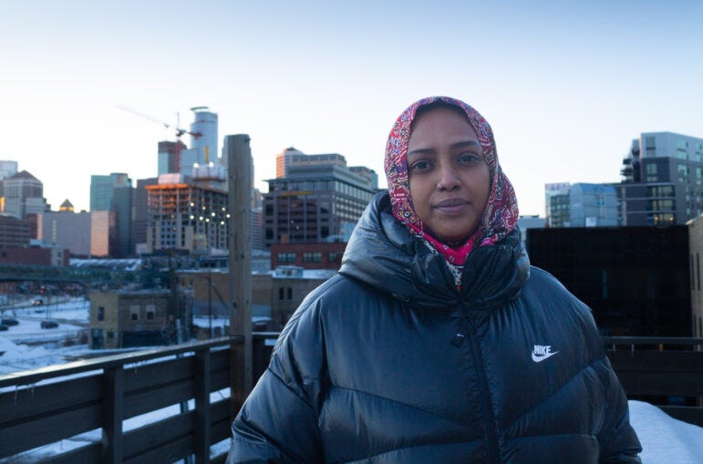 Khali Jama, dressed in a black nike puffer jacket and a red paisley hijab, stands before the downtown Minneapolis skyline. 