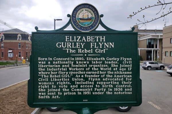 A marker consisting of a green placard with white lettering, and bearing the seal of the State of New Hampshire, honors Elizabeth Gurley Flynn, “The Rebel Girl,” making reference to her birthplace in Concord in 1890 and to her work as a nationally known labor leader. It’s pictured near a parking lot in the place where her childhood home is believed to have once stood.