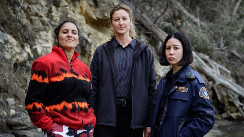 Three women stand in front of a rocky outcropping. A brunette wears brightly colored hunting clothes and stands next to a blond woman and a black-haired woman wearing police clothing