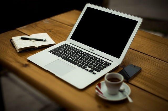 A wooden desktop holds a laptop computer, an open diary with a pen lying across it, and an espresso cup on a saucer with a spoon