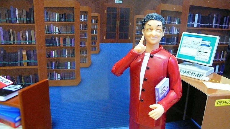 Life-like photograph of the Nancy Pearl action figure standing at the reference desk computer workstation with the book truck nearby.