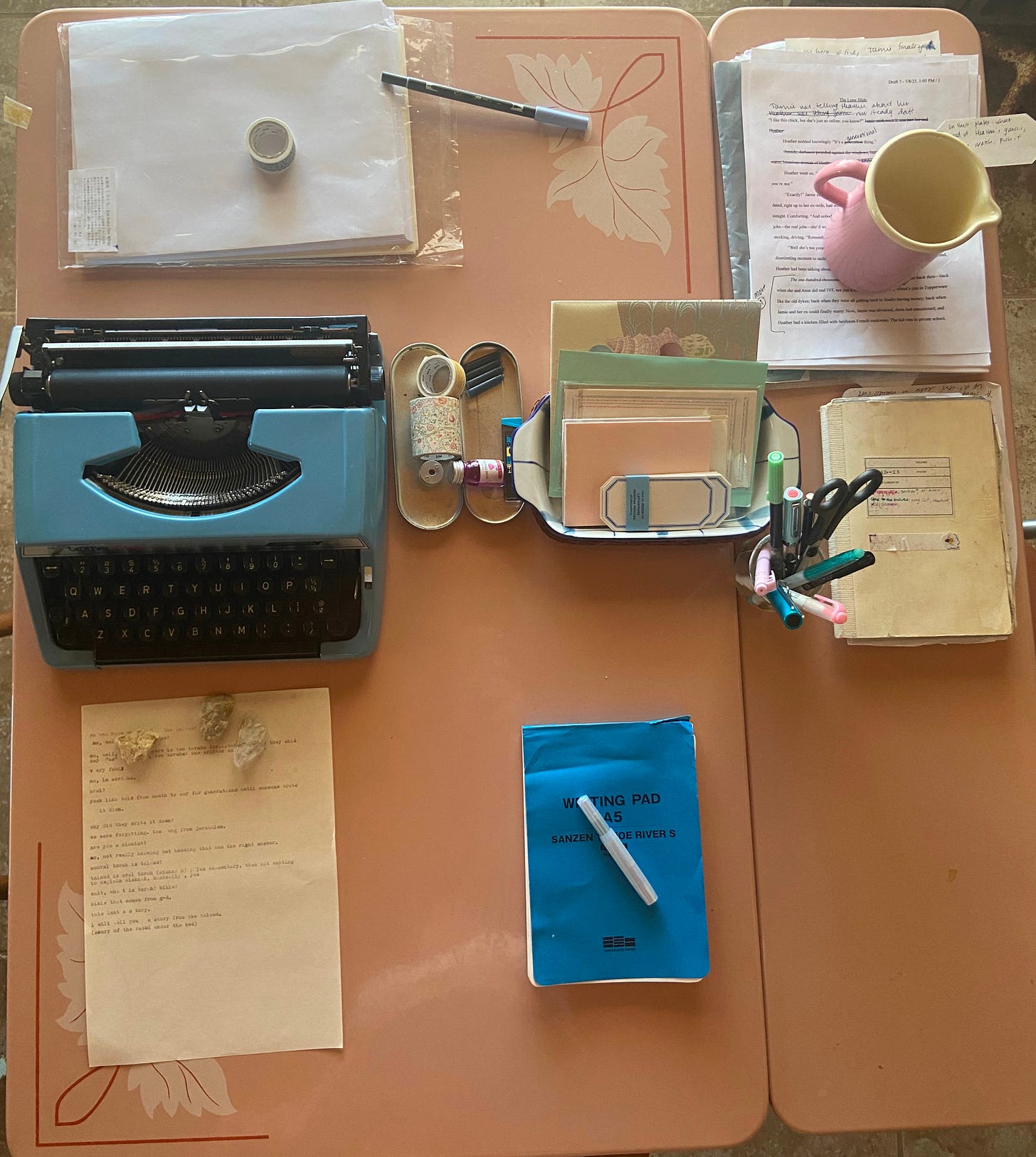 Overhead shot of a desk crammed with writing instruments: typewriter, paper, pens, ink, washi tape, and a journal.