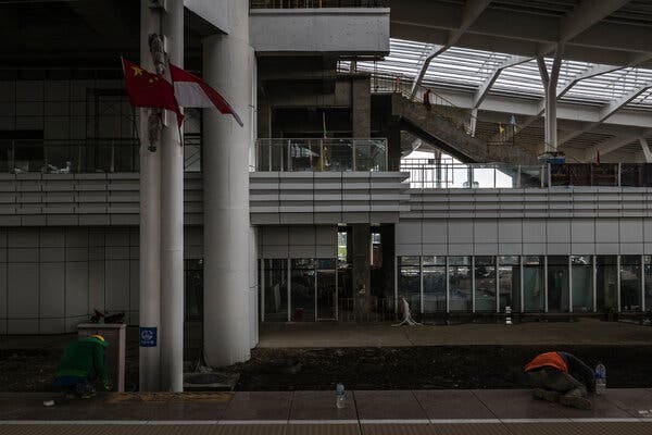Chinese and Indonesian flags hang from a column at an unfinished train station as two people on the ground work.