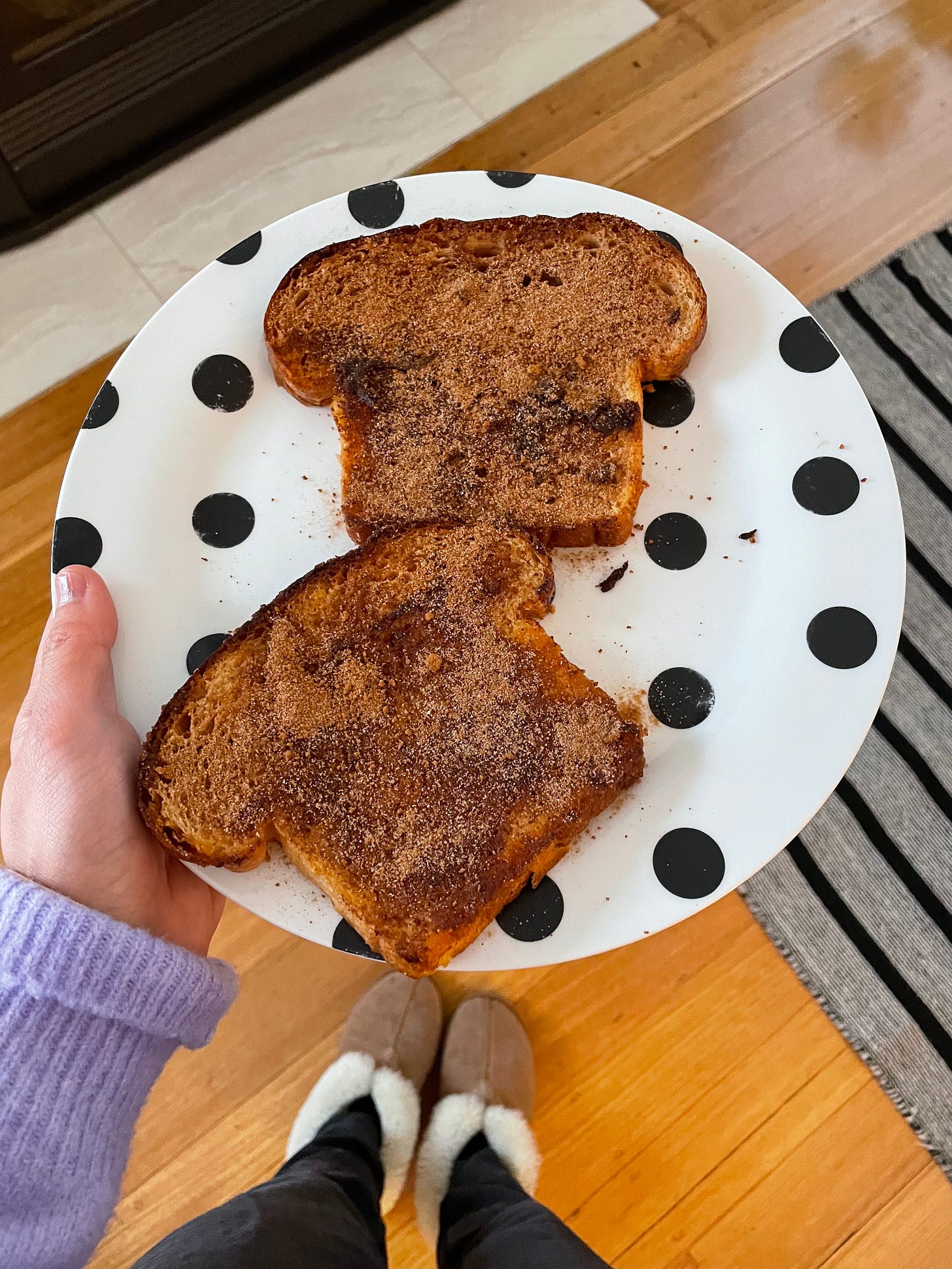 Spotty plate with two slices of milk bread which have been made into cinnamon toast. It's being held by a woman wearing a purple cardigan, track pants and ugg boots at home in winter.