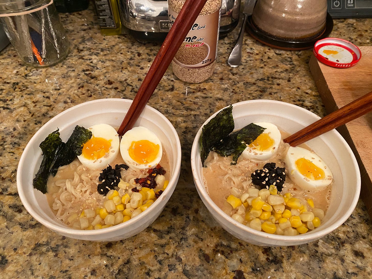 two white bowls of ramen with dark wooden chopsticks sticking out. The top of the ramen has corn, a boiled egg sliced in half, a little pile of black garlic in the centre, and sheets of gim at the side. Sesame seeds and chili crisp garnish the top, and the sesame seed container is visible on the counter in the background.