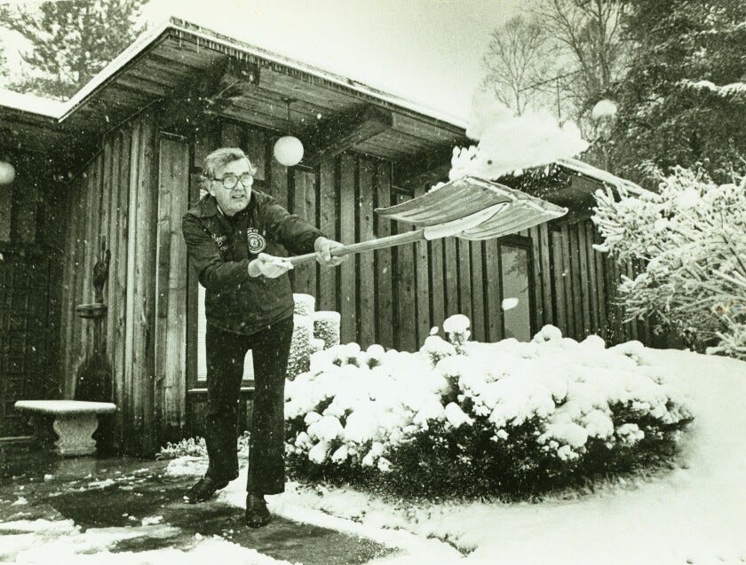 Man wearing union jacket shovels snow vigorously outside a cabin-style home.