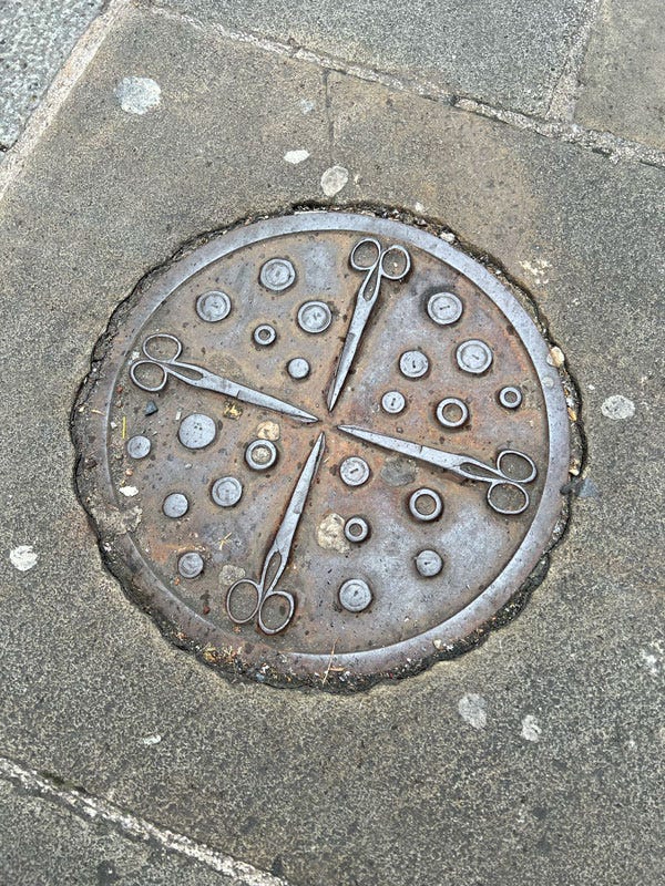 A photograph of a sidewalk manhole, made of metal, decorated with 4 scissors and what appears to be metal buttons.