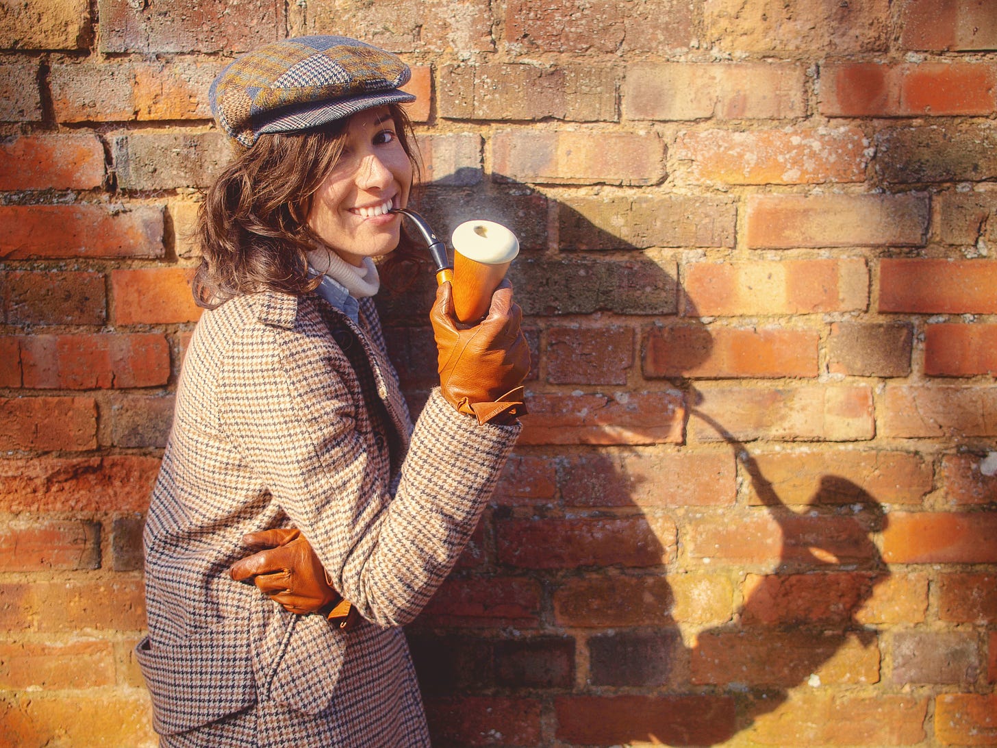 A woman in a deerstalker cap, checked coat with a pipe stands in front of a brick wall