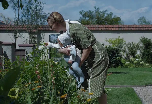 A blonde woman holds a baby while leaning over a flower garden. A fence looms in the background.