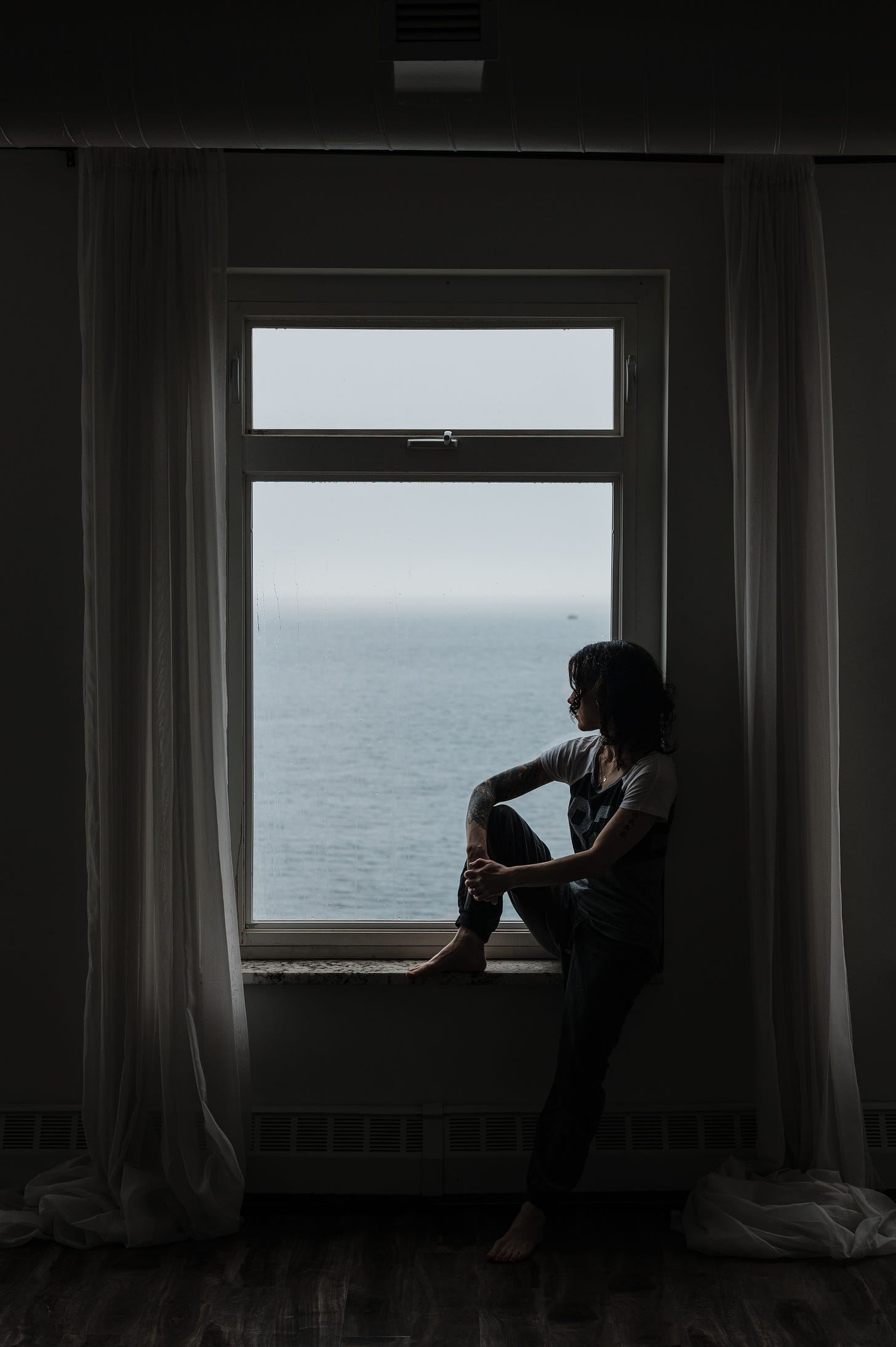author seated on the sill of a large window looking across the water in Nova Scotia