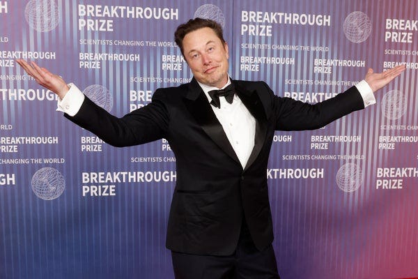 Elon Musk at the Breakthrough Prize event, dressed in a black tuxedo with arms outstretched, standing in front of the event’s branded backdrop. The Breakthrough Prize honors scientists changing the world.