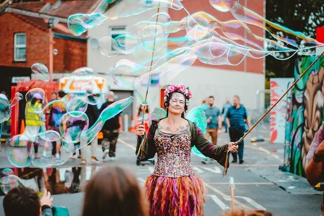 a person wearing a pink flower crown and pinkish-orange dress while holding two large bubble wands. a person wearing a pink flower crown and pinkish-orange dress while holding two large bubble wands.