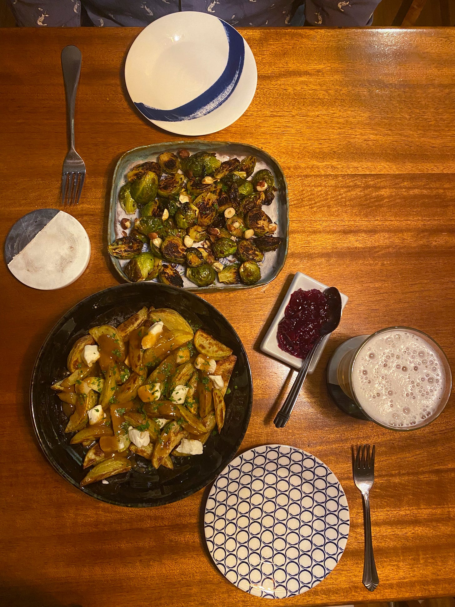From above, a black shallow dish of poutine sprinkled with parsley and an irregular bluish dish of brussels sprouts, nicely browned with hazelnuts over top. Beside them is a ramekin of cranberry sauce with a spoon in it. The dishes are arranged between two small plates with forks next to them, and a glass of beer is nearest the plate at the bottom of the frame.
